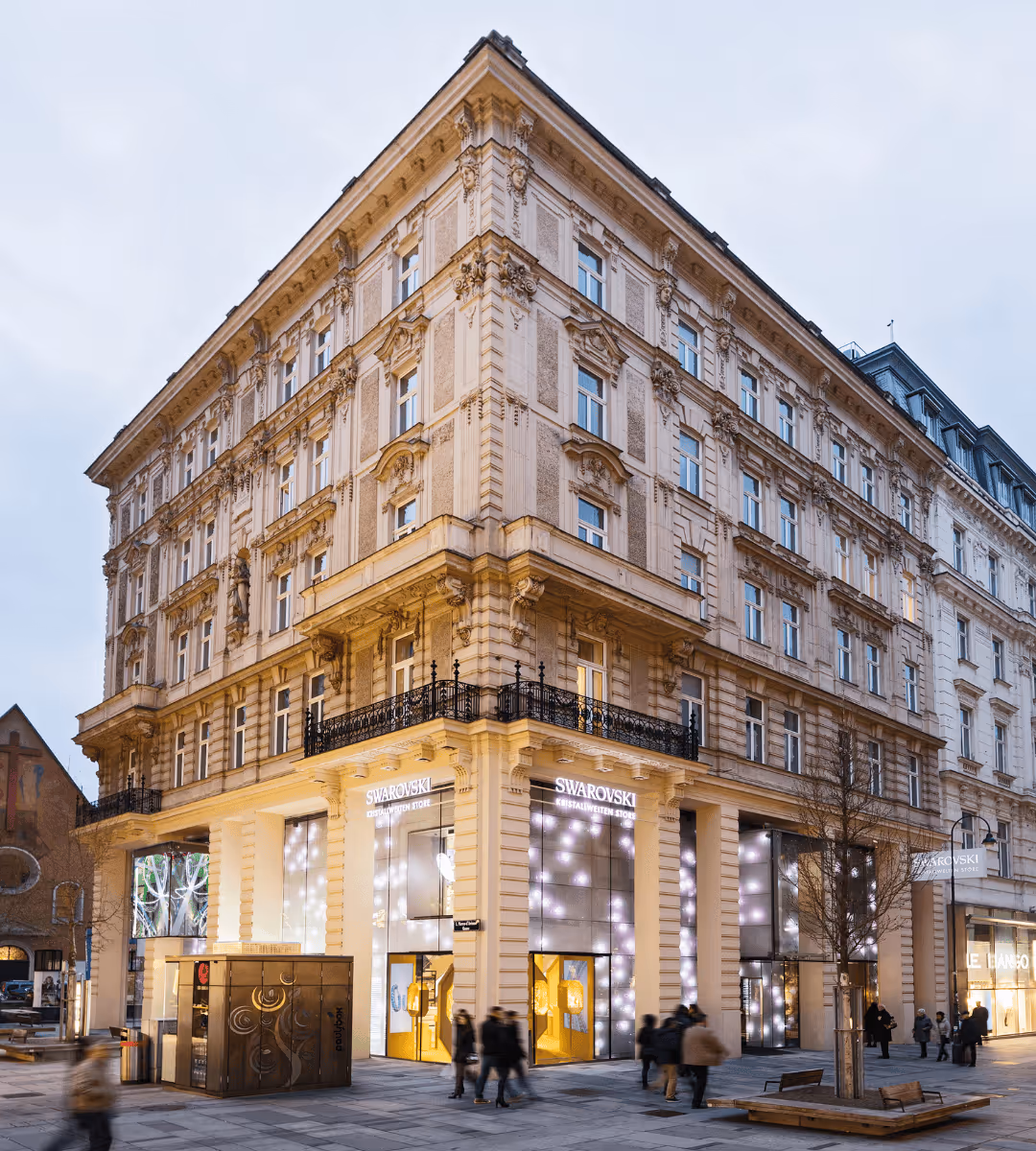Außenfassade des Swarovski Kristallwelten Stores Wien: historisches Gebäude in der Kärntner Straße mit hell erleuchteten Schaufenstern und vorbeigehenden Passanten in der Abenddämmerung.