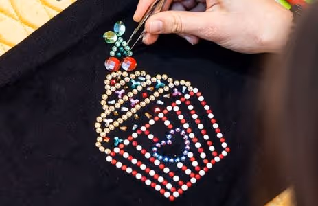 Hand sets sparkling Swarovski crystals to a cupcake motif on black fabric — birthday program “My Crystal Outfit” at Swarovski Kristallwelten.