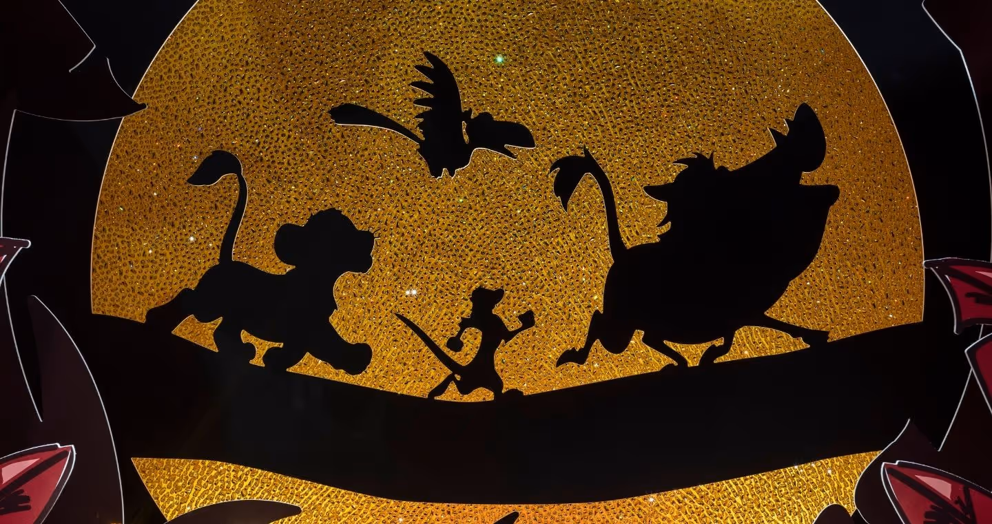 Disney Der König der Löwen Kunstbühne im Swarovski Kristallwelten Store Wattens – schwarze Silhouetten von Simba, Timon, Pumbaa und Zazu auf einem Ast vor einer goldfunkelnden Sonne, umrahmt von roten Blättern.