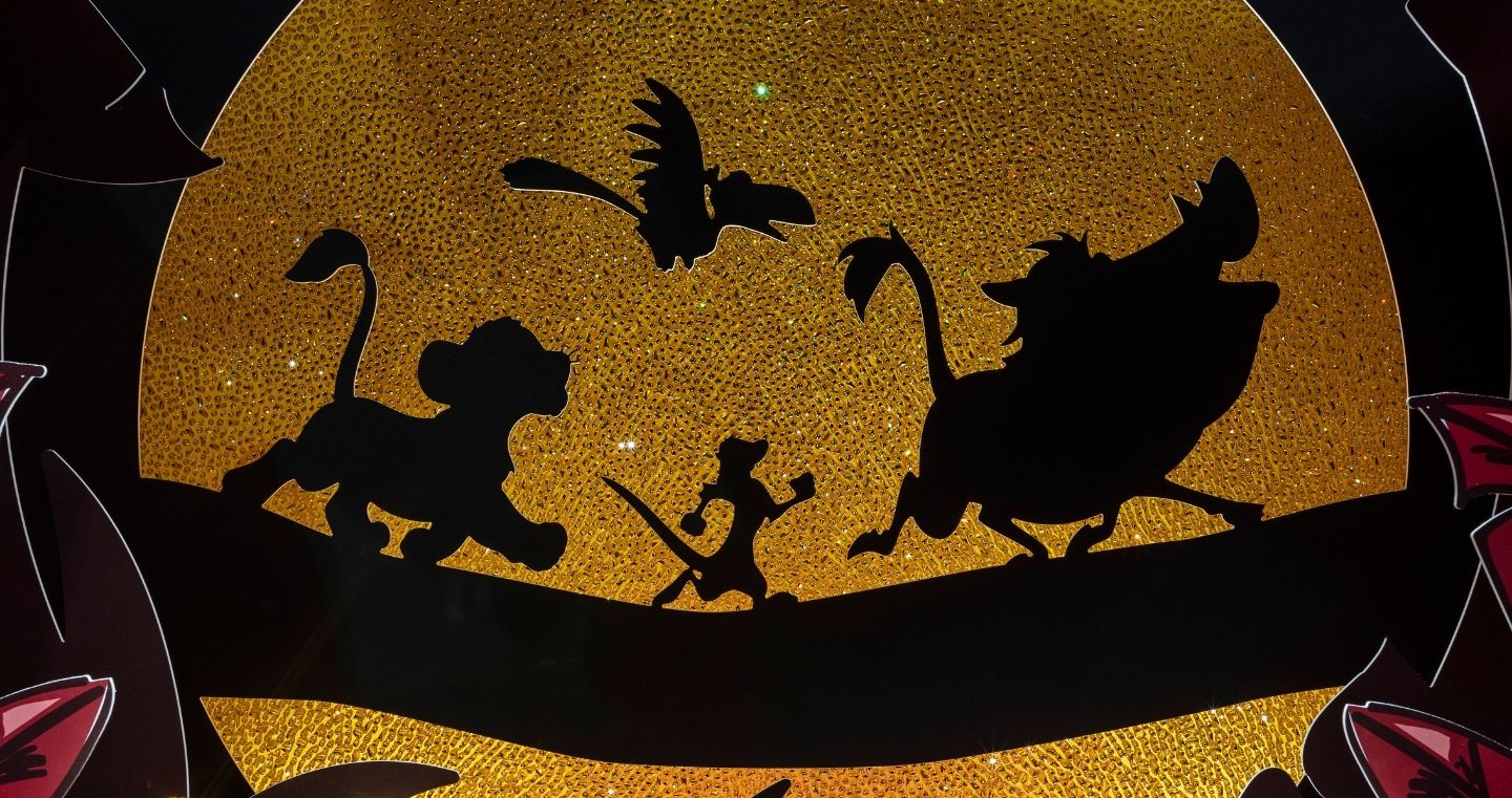 Disney The Lion King art stage at Swarovski Kristallwelten Store Wattens — black silhouettes of Simba, Timon, Pumbaa and Zazu on a branch in front of a sparkling golden sun, framed by red leaves.