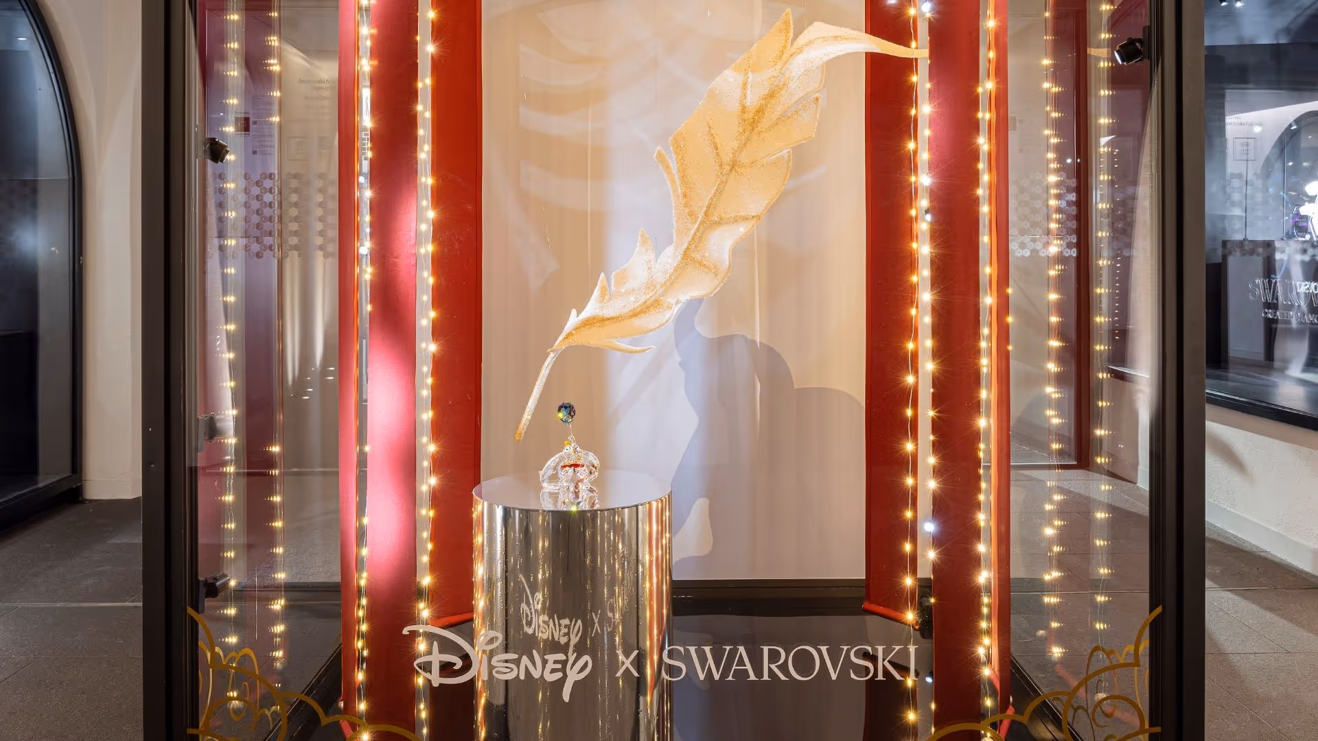 Disney x Swarovski Kunstbühne im Swarovski Kristallwelten Store Innsbruck – große goldene Kristallfeder schwebt über eine Dumbo Kristallfigur, umrahmt von roten Vorhängen und Lichterketten.