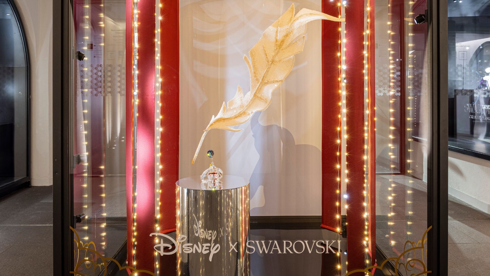 Disney x Swarovski Kunstbühne im Swarovski Kristallwelten Store Innsbruck – große goldene Kristallfeder schwebt über eine Dumbo Kristallfigur, umrahmt von roten Vorhängen und Lichterketten.