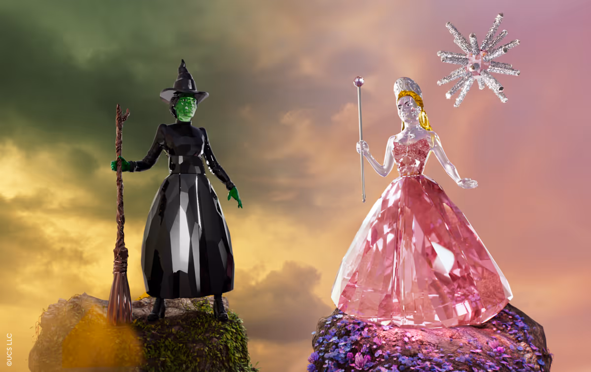 Zwei Kristallfiguren aus der Wicked x Swarovski Kollektion: die grüne Hexe in Schwarz und Glinda in Rosa stehen sich auf funkelnden Felsen gegenüber, vor einem farbenprächtigen Himmel.