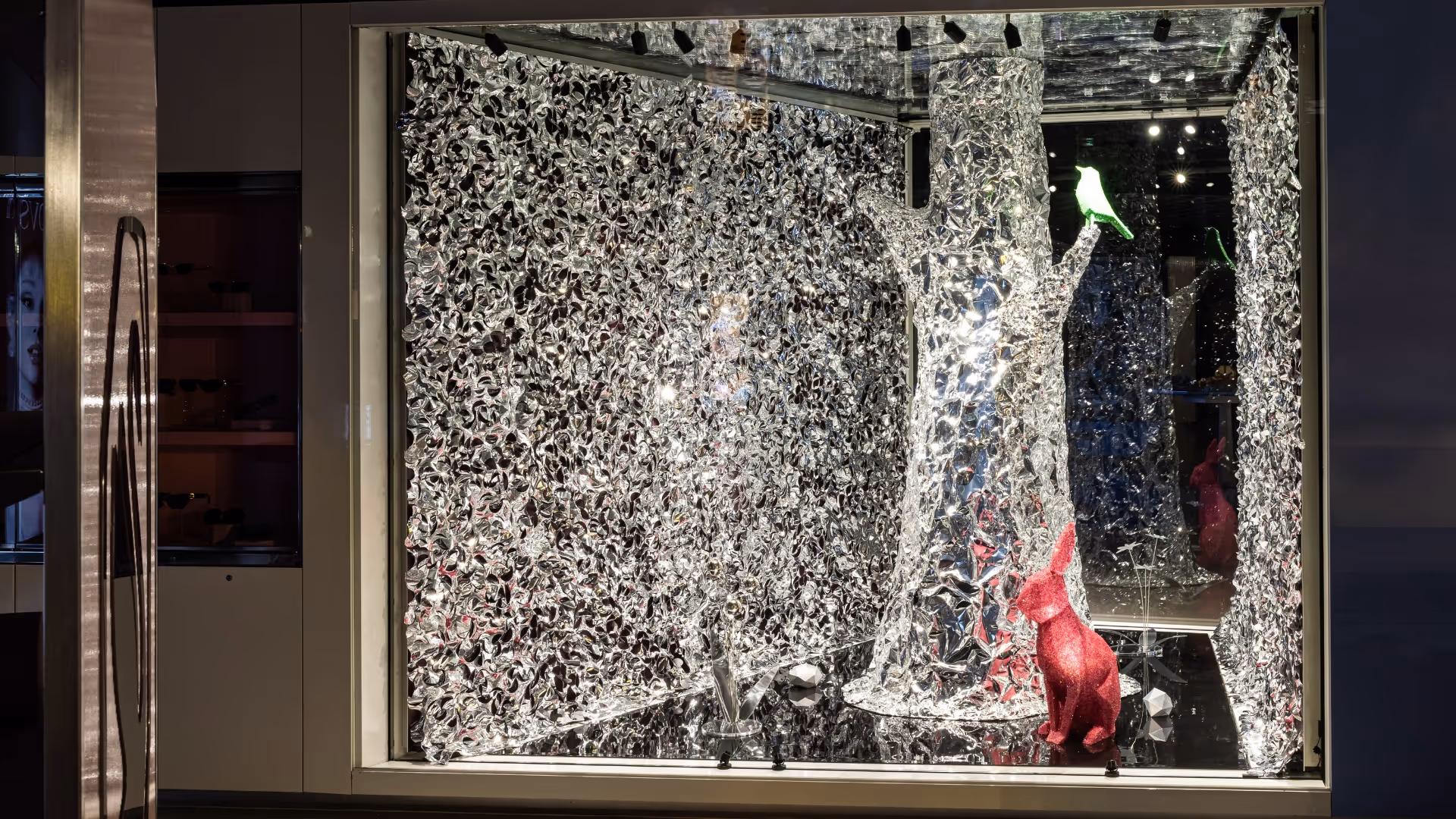 In the second showcase, a faceted metallic tree supports a bucolic green faceted crystal bird. On the floor a pink crystal rabbit is surrounded by mirrored flowers. This decor is surrounded by faceted metallic wall and floors.