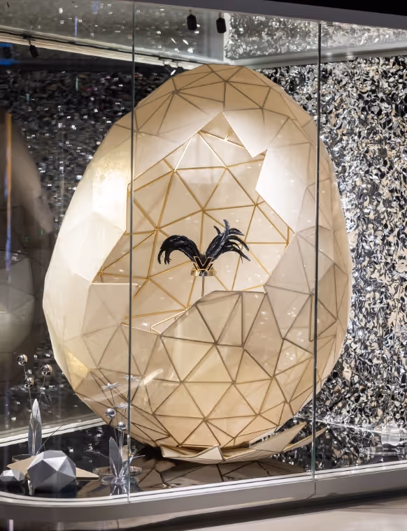 An immense faceted crystal egg is standing in the middle of a showcase inside of a store. It is partly cracked. The broken fragments stand on the floor. Inside the egg, there is a headpiece from the Swarovski Corporate Archive with feathers.