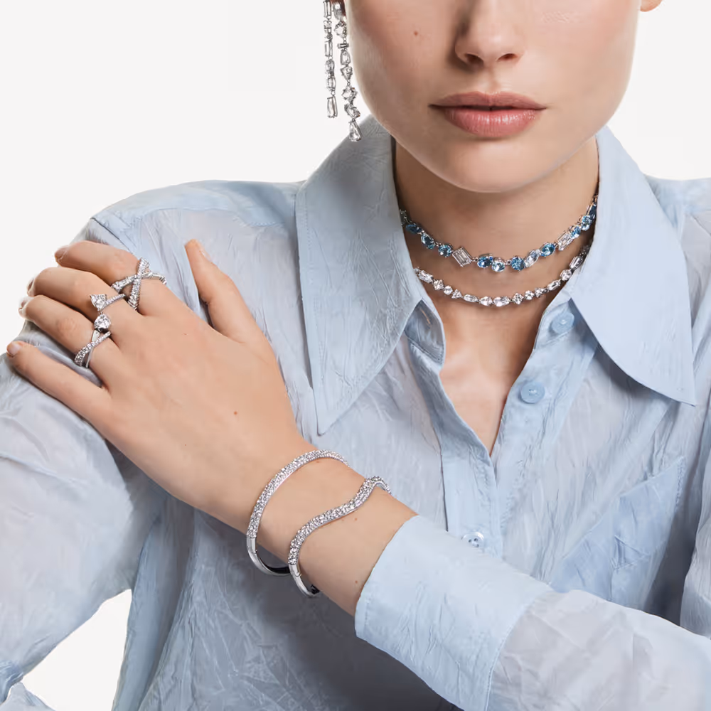 Model wears elegant Swarovski silver jewelry with clear crystals: two filigree necklaces, sparkling bangles, multiple rings and long earrings. The jewelry is styled into a light blue blouse and looks modern, cool and luxurious. Focus on neck, wrist, and hand details against a neutral background.