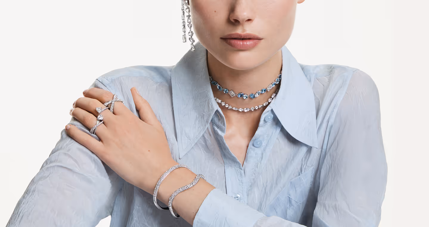 Model wears elegant Swarovski silver jewelry with clear crystals: two filigree necklaces, sparkling bangles, multiple rings and long earrings. The jewelry is styled into a light blue blouse and looks modern, cool and luxurious. Focus on neck, wrist, and hand details against a neutral background.