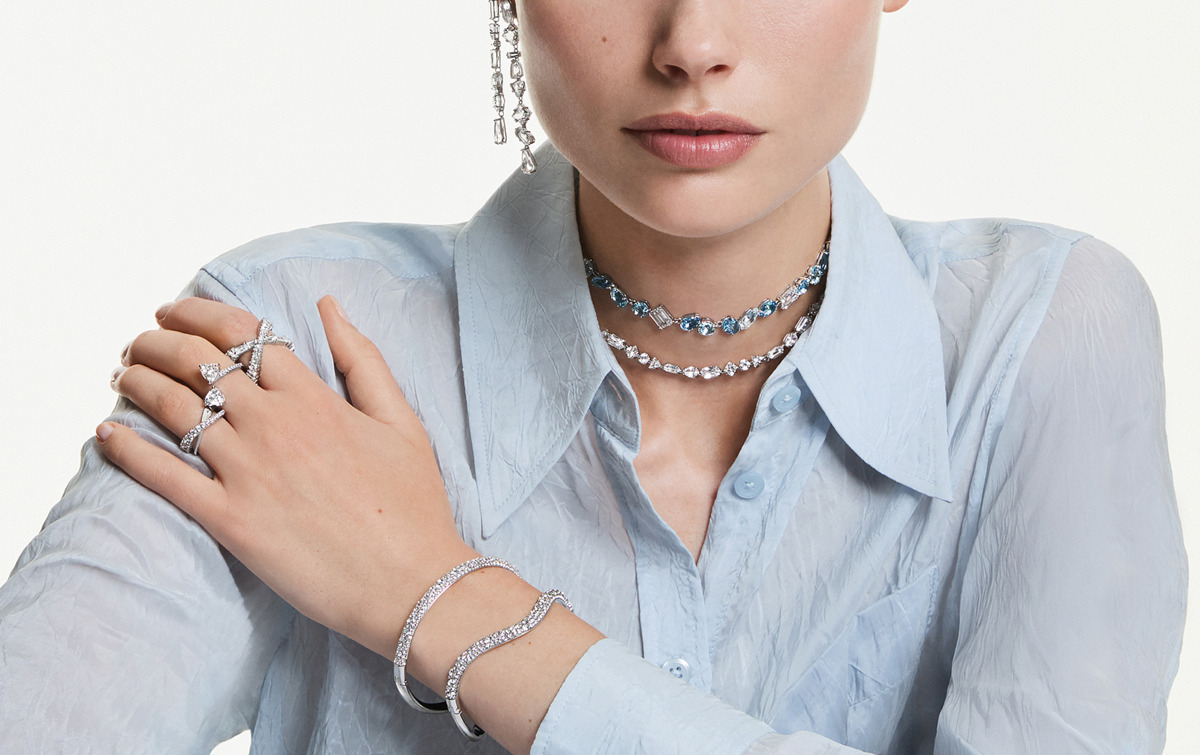 Model trägt eleganten Swarovski Silberschmuck mit klaren Kristallen: zwei filigrane Halsketten, funkelnde Armreifen, mehrere Ringe und lange Ohrringe. Der Schmuck wird zu einer hellblauen Bluse gestylt und wirkt modern, kühl und luxuriös. Fokus auf Details von Hals, Handgelenk und Hand vor neutralem Hintergrund.