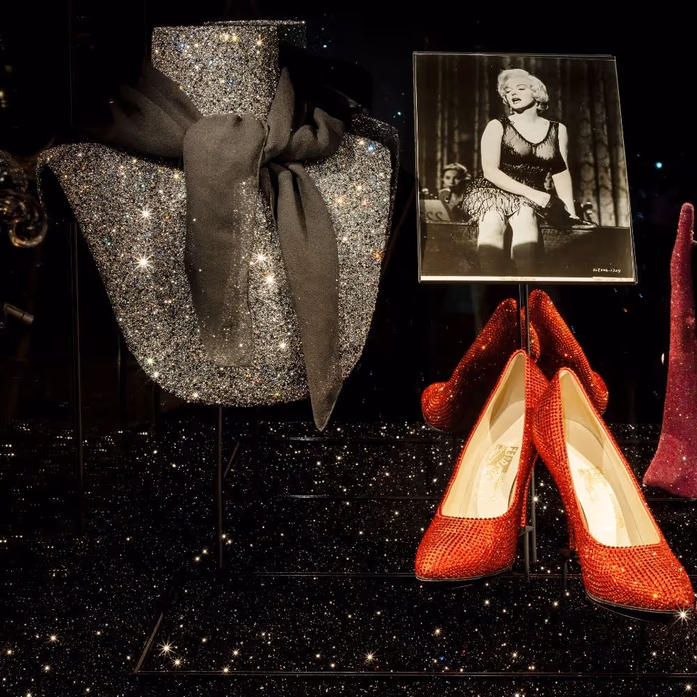 Eine Ausstellungspräsentation zeigt einen mit Kristallen bedeckten Torso auf einem schwarzen Sockel, verziert mit einer großen schwarzen Stoffschleife. Daneben steht ein Schwarz-Weiß-Foto, das eine Bühnenszene mit einer Person in einem funkelnden Bühnenkostüm zeigt. Im Vordergrund befindet sich ein Paar leuchtend rote, kristallbesetzte High Heels auf einer schwarzen Fläche. 