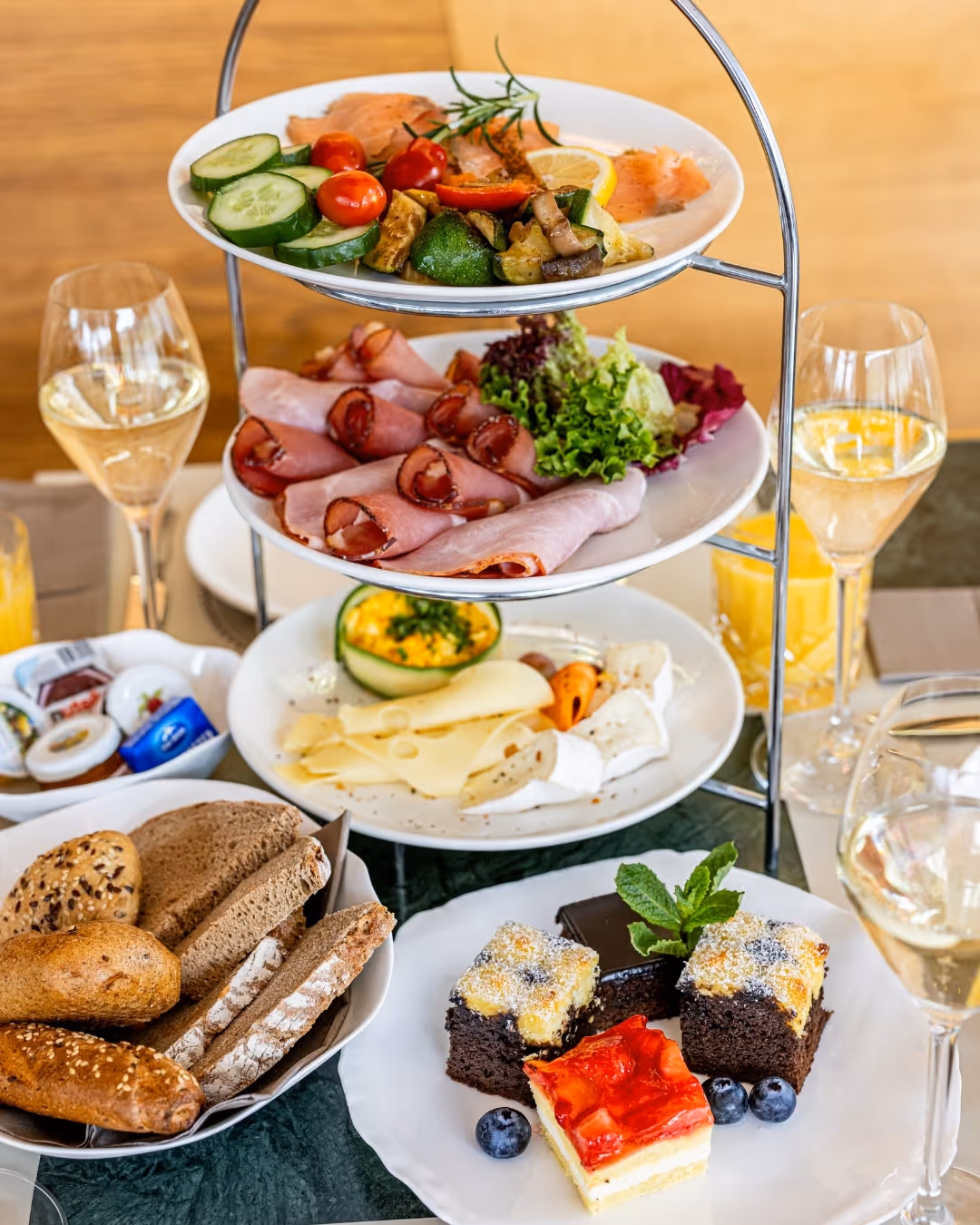 Etagere with brunch: salmon, ham, cheese, vegetables; plus bread, cake and drinks such as juice and sparkling wine on a set table.