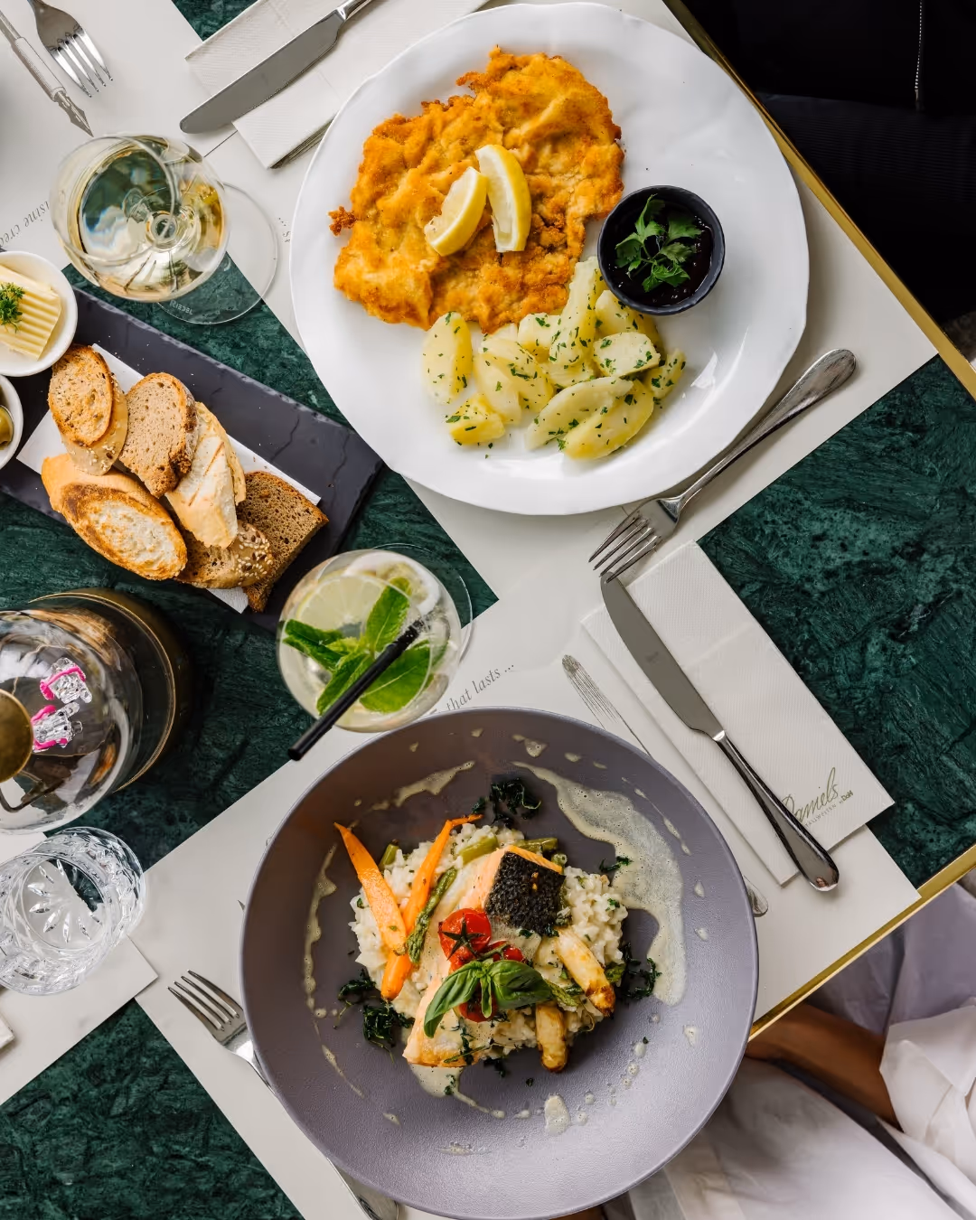 Teller mit Wiener Schnitzel, Petersilienkartoffeln, Beerenkompott sowie Risotto mit Fisch; dazu Brot, Butter, Getränke und Besteck auf grünem Marmortisch.