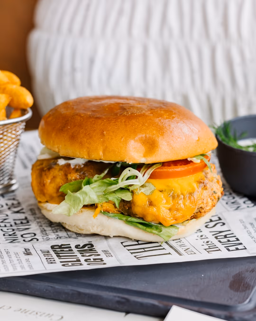 Juicy cheeseburger with lettuce and tomato in a brioche roll, served on paper, with fries and dip in the background.