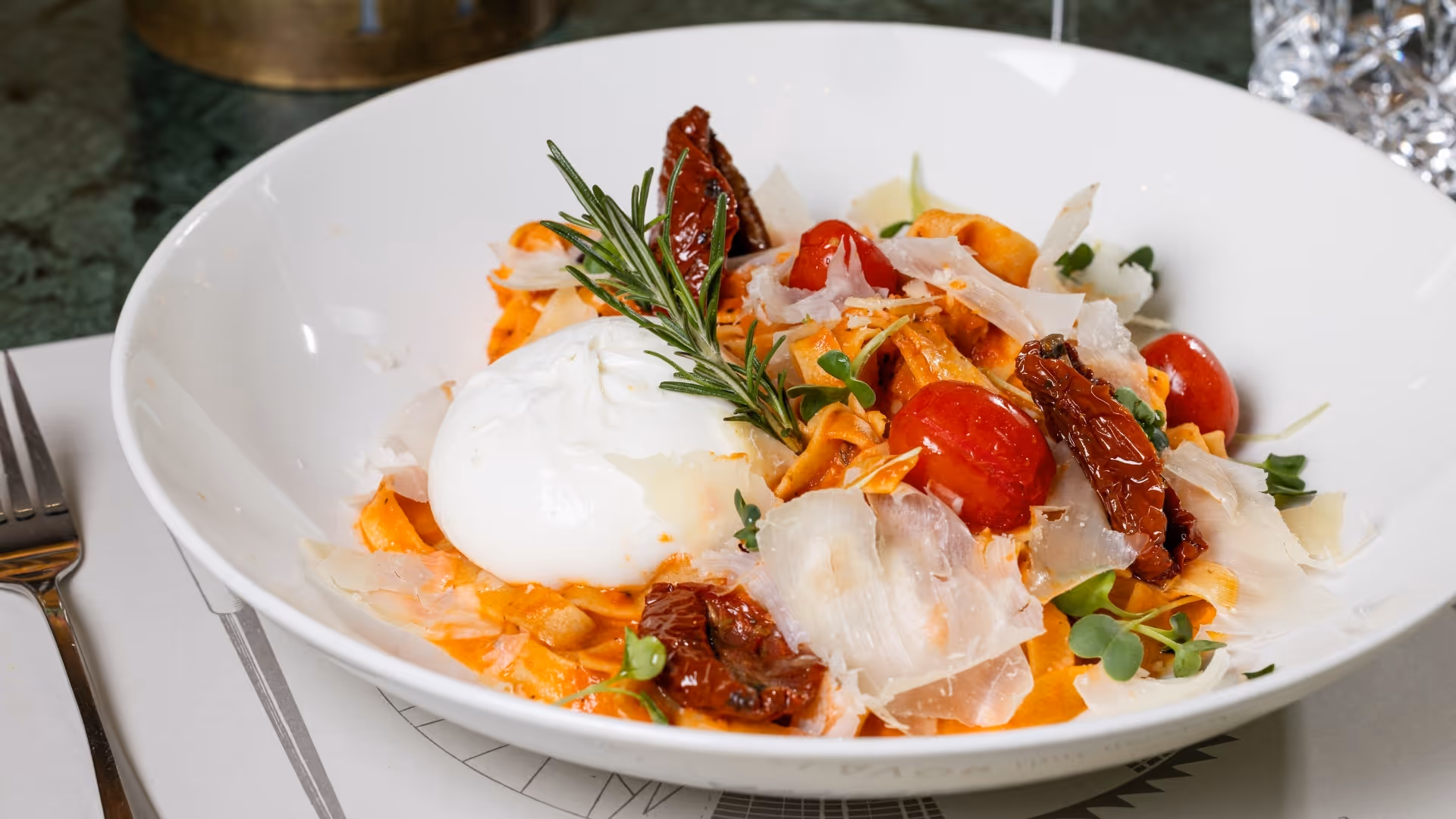 Pasta with creamy burrata, cherry tomatoes, sun-dried tomatoes, parmesan and herbs, served in a white bowl on a set table.