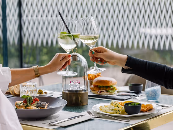 Two people toast at a set table with wine glasses; burgers, schnitzel with potatoes, fish on risotto, bread and side dishes are served.