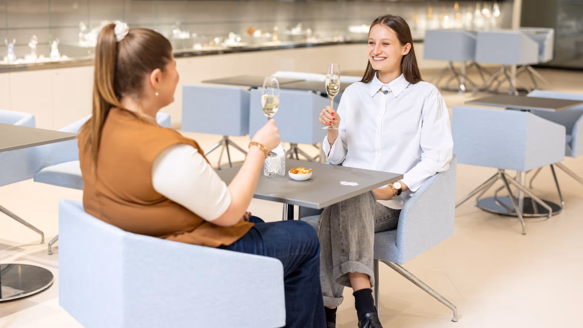 Zwei Personen stoßen mit Weißweingläsern an einem modern gedeckten Tisch an; im Hintergrund helle Lounge mit Sitzmöbeln und Auslage.