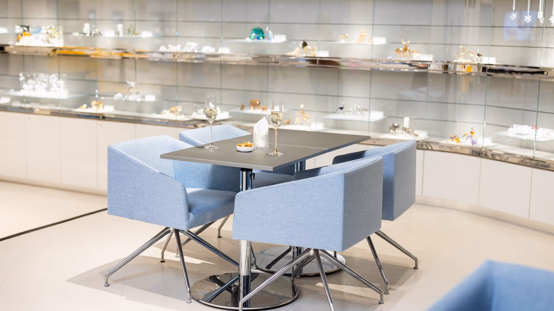 Moderner Tisch mit zwei Stühlen und Gläsern Weißwein; dahinter beleuchtete Vitrinen mit ausgestellten Kristallobjekten in heller Lounge-Umgebung.