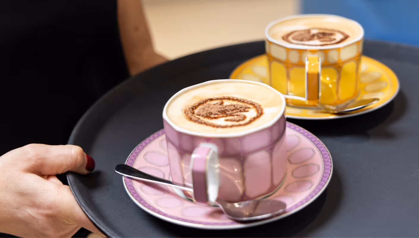 Zwei Cappuccino-Tassen auf Tablett, eine rosa und eine gelbe, mit Milchschaum-Muster, serviert mit Löffeln auf Untertassen.