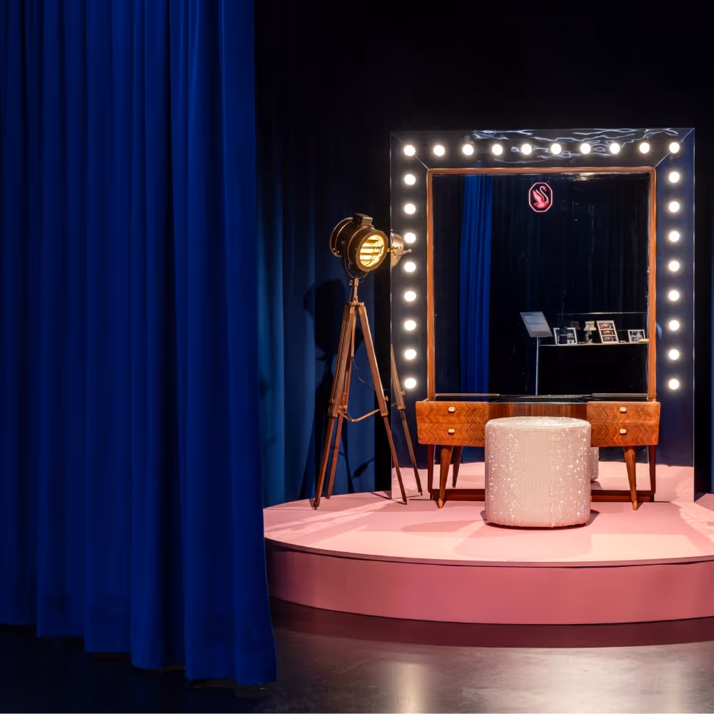 Eleganter Schminkplatz auf rosa Bühne mit beleuchtetem Spiegel, funkelndem Hocker und Studiolampe, eingerahmt von dunkelblauen Vorhängen.