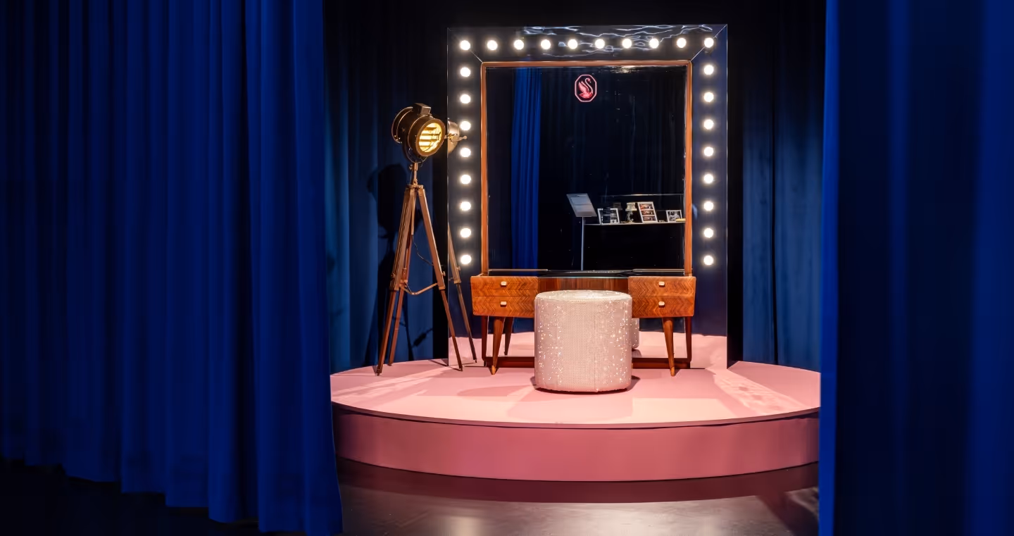 Elegant makeup space on a pink stage with illuminated mirror, sparkling stool and studio lamp, framed by dark blue curtains.