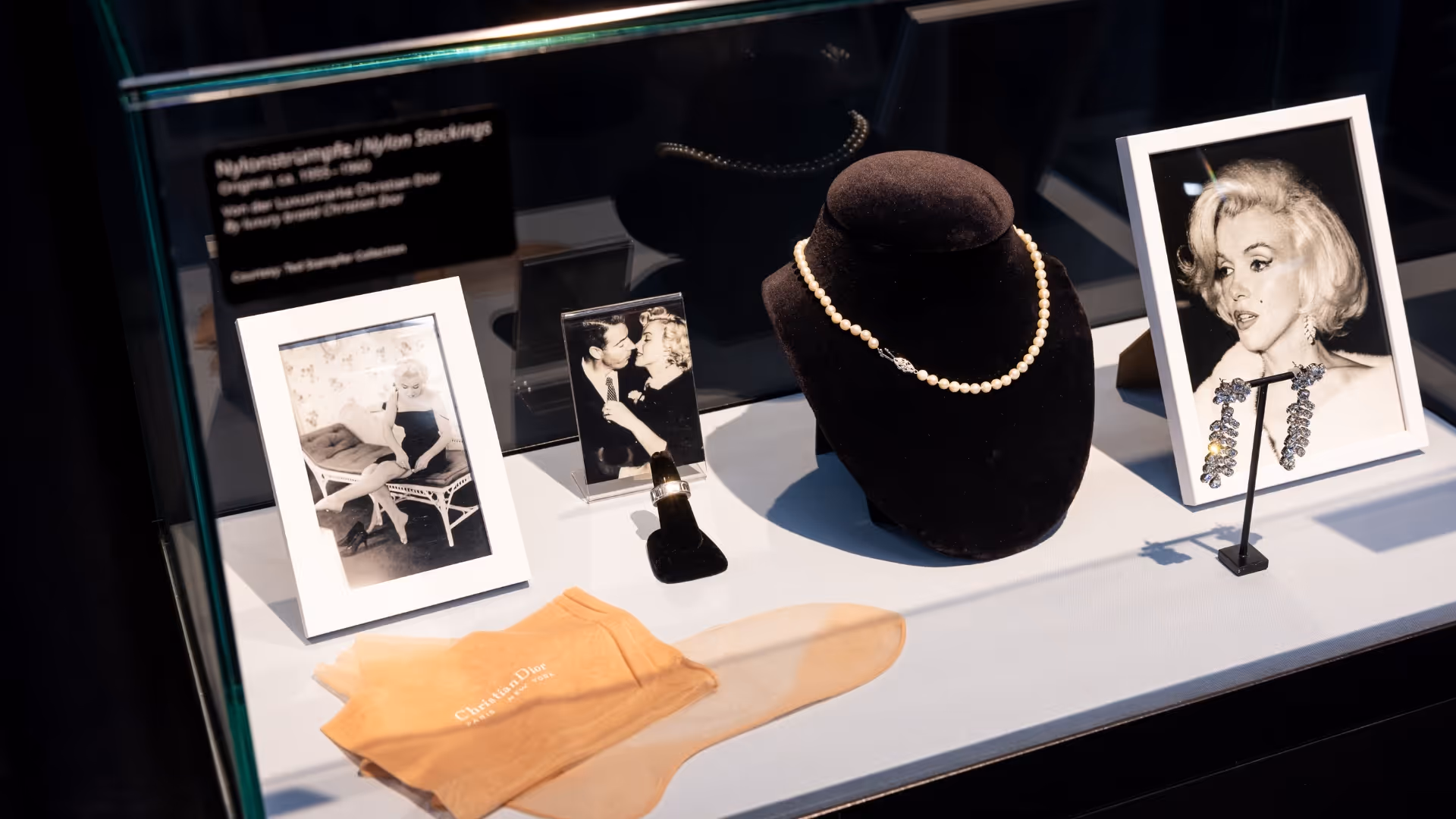 Vitrine mit Dior‑Nylonstrümpfen, Perlenkette auf schwarzem Display, Ring, glitzernden Ohrringen und gerahmten Schwarz‑Weiß‑Fotos.