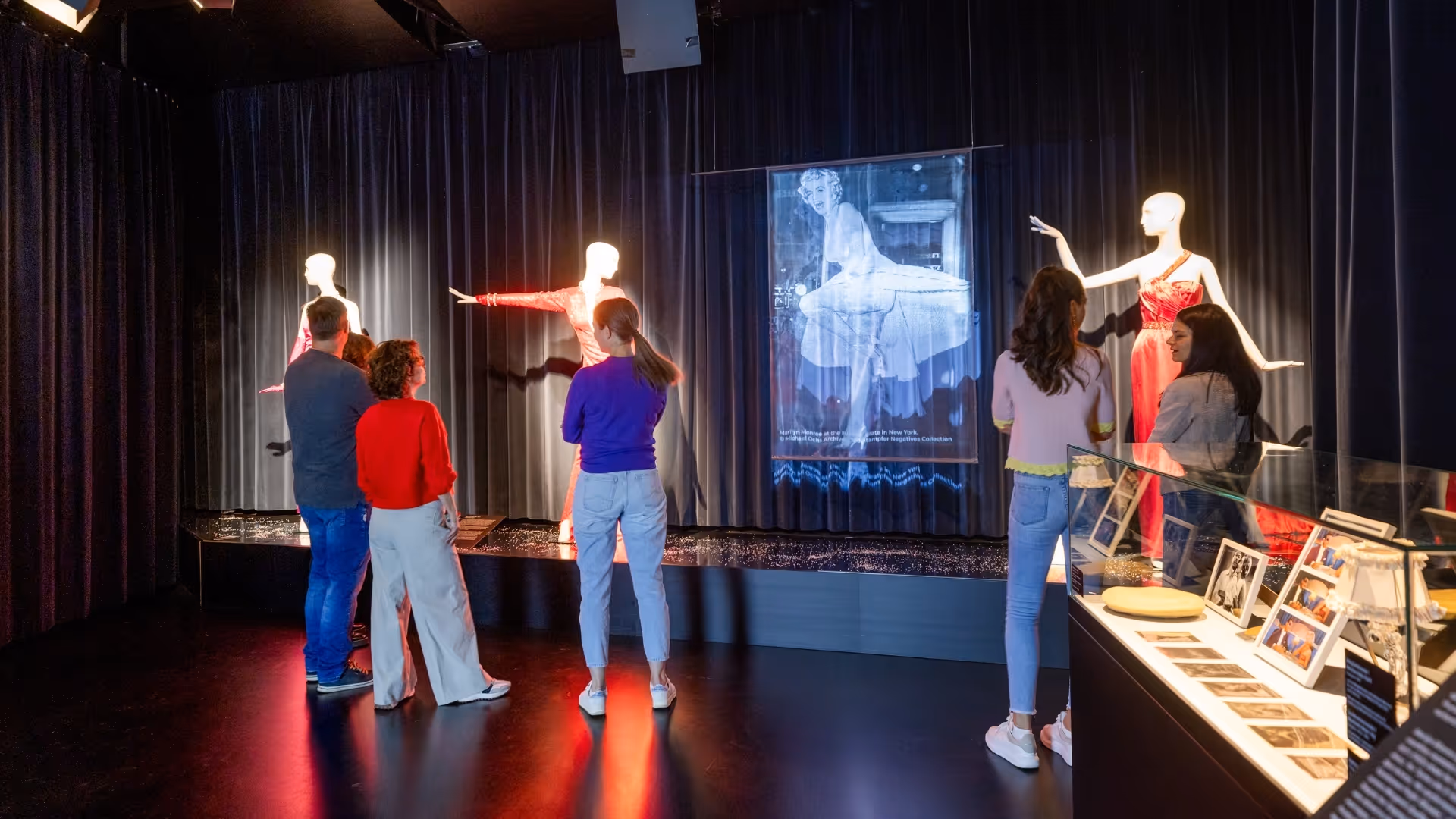 Mehrere Besucher*innen betrachten eine Ausstellung mit Mannequins in roten Kleidern und einer Projektion, daneben eine Vitrine mit Fotos und Objekten.
