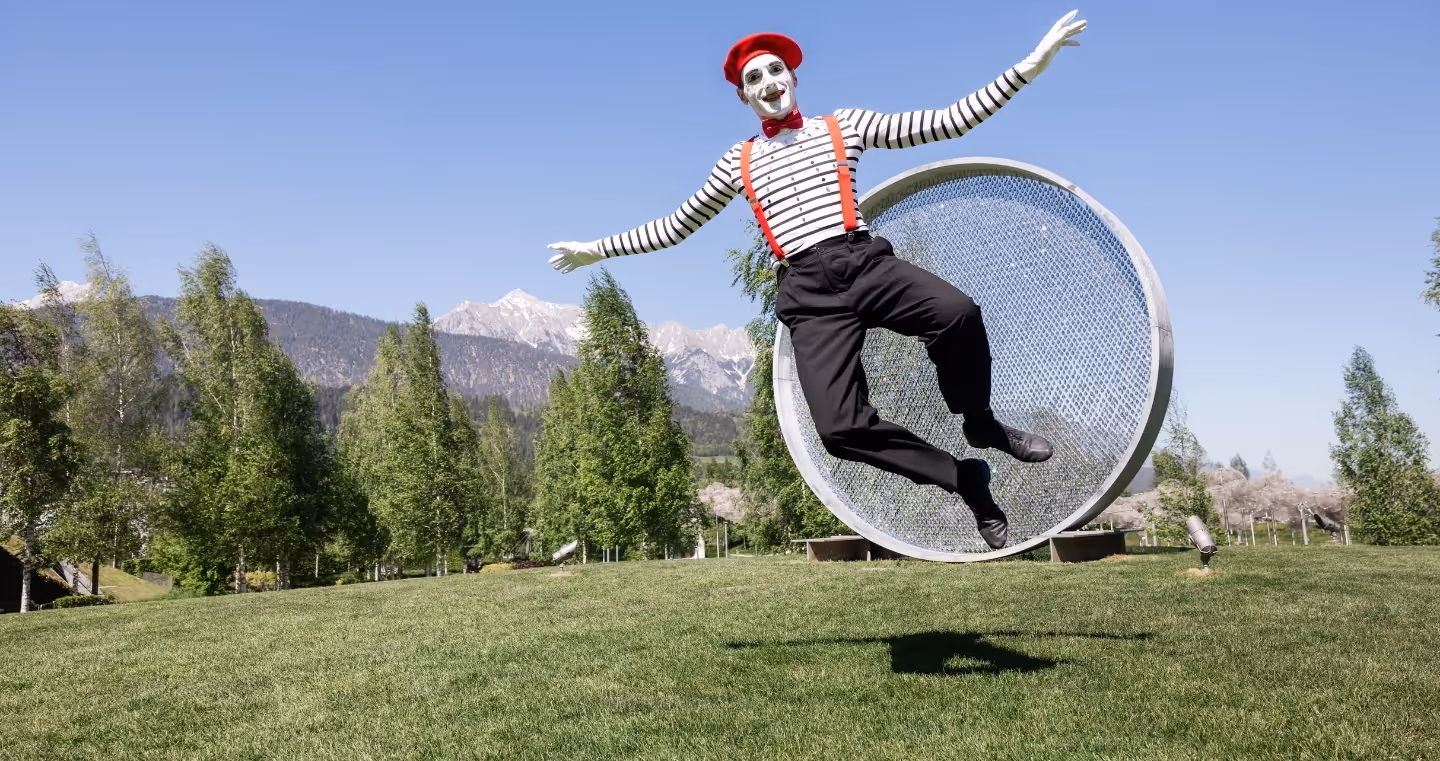 Pantomime in gestreiftem Shirt, roter Mütze und Hosenträgern springt im Park der Swarovski Kristallwelten mit Bäumen und Bergen im Hintergrund.