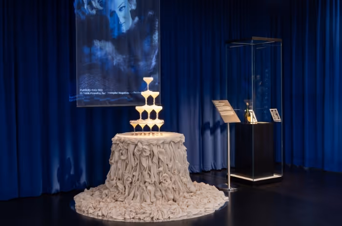 Installation mit Champagnerturm auf rundem Tisch, daneben eine Vitrine; im Hintergrund Projektion eines Porträts von Marilyn Monroe vor blauen Vorhängen.