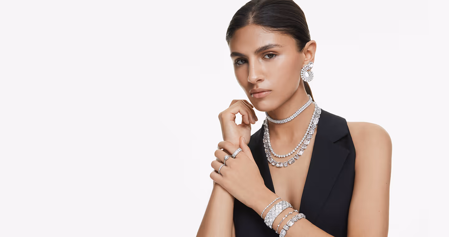 Person mit zurückgebundenem Haar trägt funkelnden Swarovski Schmuck: Halsketten, Ohrringe, Ringe und Armbänder, kombiniert mit schwarzem Outfit vor hellem Hintergrund.
