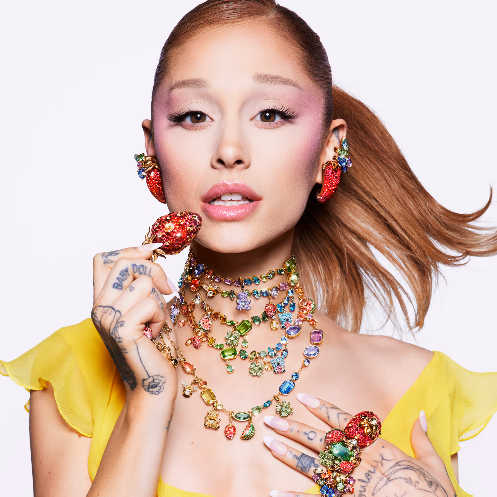 A person with a ponytail wears a yellow top and eye-catching, colorful Swarovski jewelry with strawberry motifs, multiple necklaces and rings against a light background.