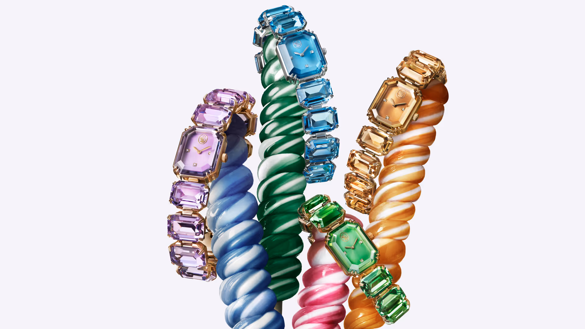 Several colorful Swarovski wristwatches with crystals in blue, green, purple and gold, arranged around twisted, candy-like decorative elements against a light background.