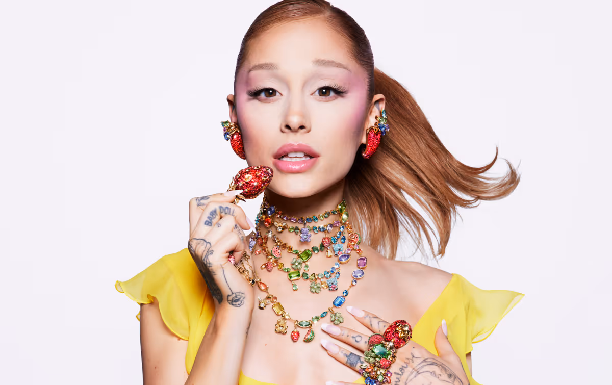 A person with a ponytail wears a yellow top and eye-catching, colorful Swarovski jewelry with strawberry motifs, multiple necklaces and rings against a light background.