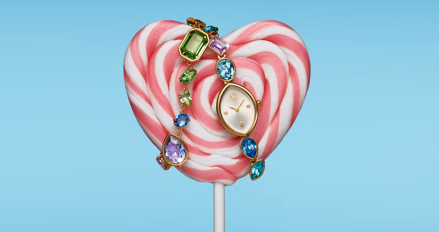 Swarovski Armbanduhr mit bunten Kristallen vor einem rosa-weißen Herz-Lolli auf hellblauem Hintergrund – verspielte Inszenierung mit Candy-Flair.