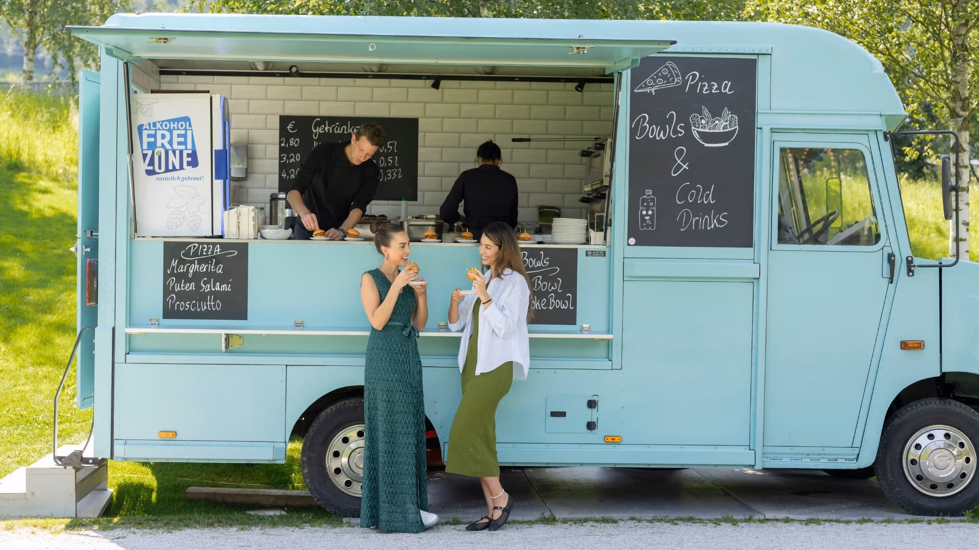 Two visitors enjoy snacks in front of a food truck with pizza, bowls and drinks in the park of Swarovski Kristallwelten.