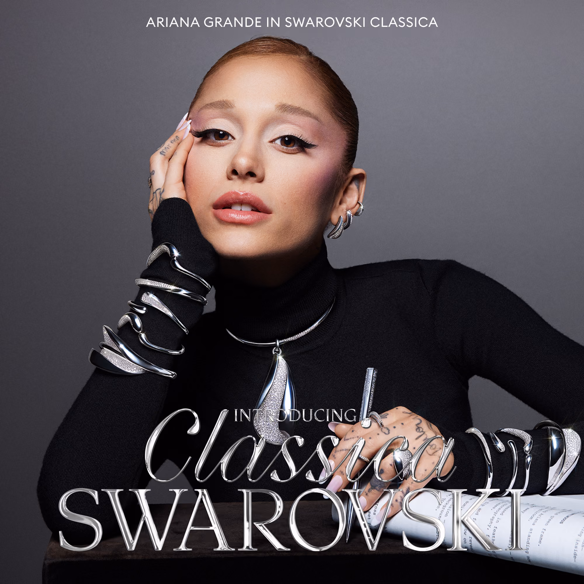 Person wearing a black turtleneck sweater with silver-colored Swarovski jewelry on arms and neck against a grey background.