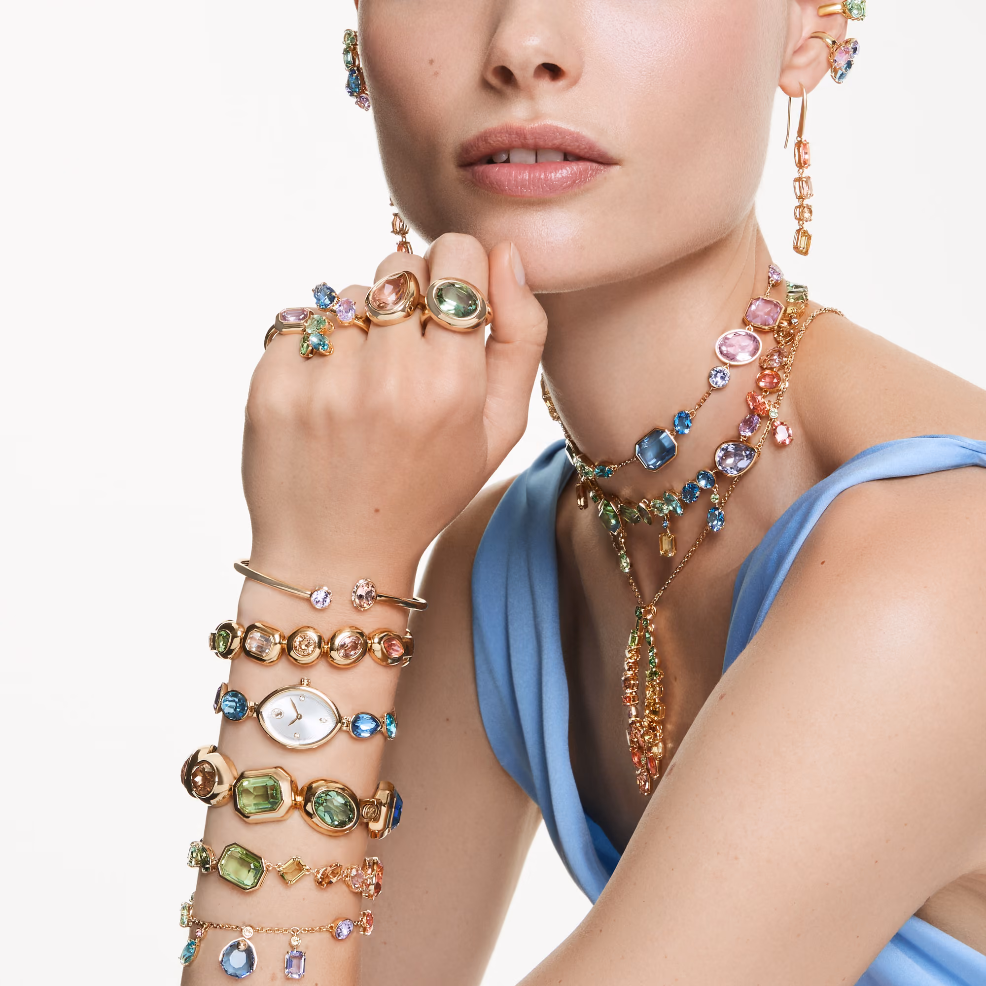 Close-up of a person wearing colorful Swarovski jewelry: rings, bracelets, watch, earrings and multi-strand necklaces with pastel colored crystals.