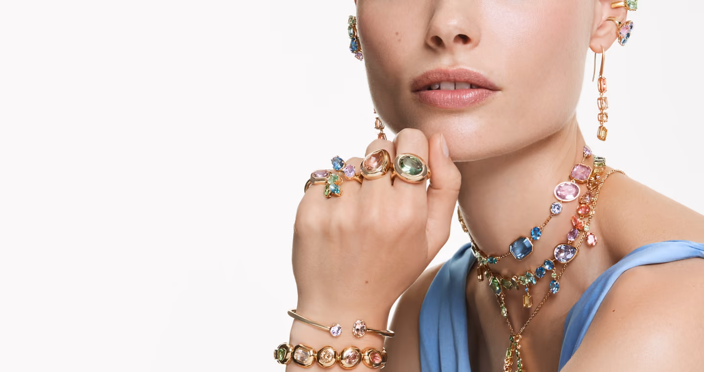 Close-up of a person wearing colorful Swarovski jewelry: rings, bracelets, watch, earrings and multi-strand necklaces with pastel colored crystals.