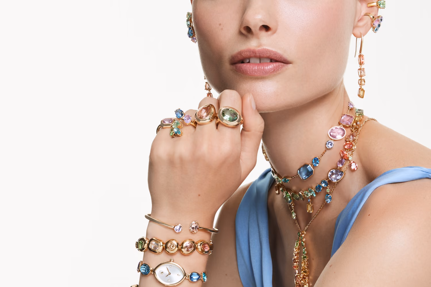 Close-up of a person wearing colorful Swarovski jewelry: rings, bracelets, watch, earrings and multi-strand necklaces with pastel colored crystals.