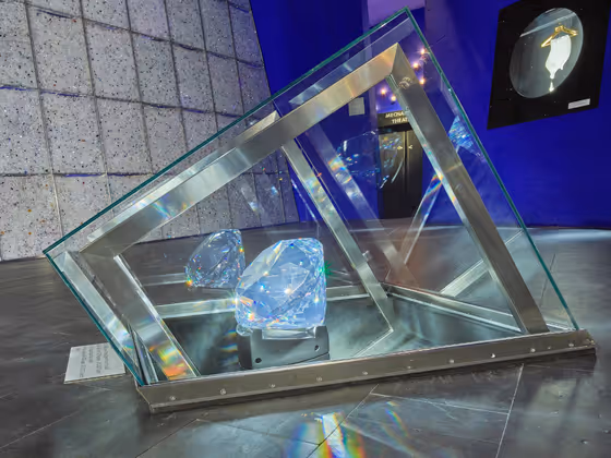 Kunstwerk von Hubert Steinlechner in den Swarovski Kristallwelten: Ein großer geschliffener Kristall liegt in einer geometrischen Metall- und Glasinstallation, die sich wie ein Rahmen um den Kristall legt. Der Hintergrund zeigt eine glitzernde Wand.