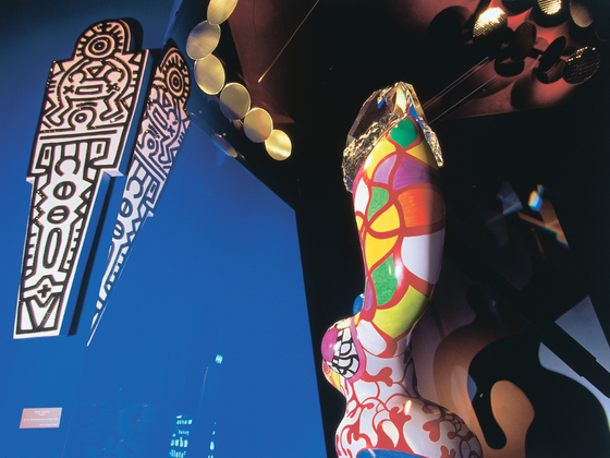 Farbintensive Skulptur von Niki de Saint Phalle: Eine bunte, organisch geformte Figur mit kräftigen Mustern in Blau, Rot, Grün und Gelb, die einen übergroßen Kristall auf einer ausgestreckten Armform trägt.
