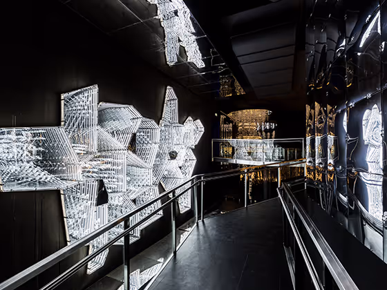 Kunstwerk 'Into Lattice Sun': Ein dunkler Raum, durch den ein Steg führt. Links und rechts davon kristalline, urbane Objekte.