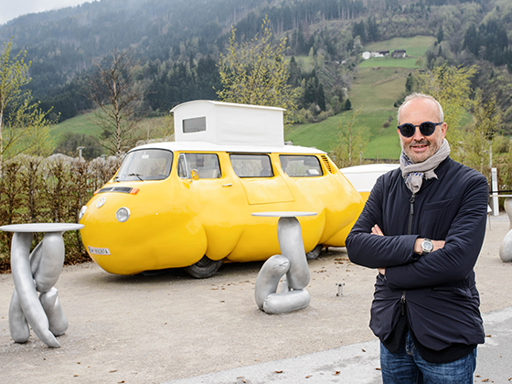 Erwin Wurm, österreichischer Künstler, 2017 mit Sonnenbrille und Schal, posierend vor seiner Installation "Dicker Bus"