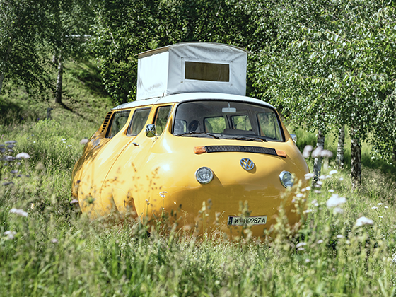 Skulptur 'Dicker Bus': Ein verzerrt wirkender, bauchiger Bus in Grau, humorvoll inszeniert.