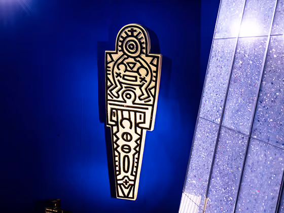 Wandinstallation in mit einem typischen Keith-Haring-Motiv: eine stilisierte, grafische Figur mit schwarzen Kristallen auf weißem Grund mit Symbolen und Mustern. Das Werk leuchtet auffällig vor der blauen Wand.