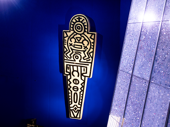 Wandinstallation in mit einem typischen Keith-Haring-Motiv: eine stilisierte, grafische Figur mit schwarzen Kristallen auf weißem Grund mit Symbolen und Mustern. Das Werk leuchtet auffällig vor der blauen Wand.