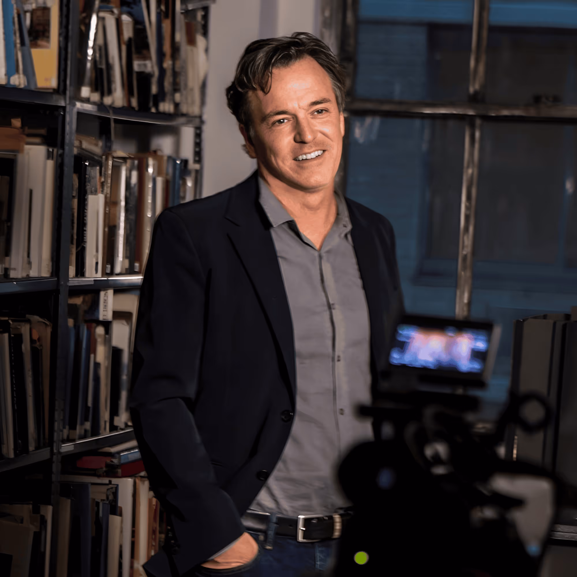 Derek McLane, US-amerikanischer Bühnenbildner, im Porträt mit schwarzer Jacke, stehend vor Bücherregalen.