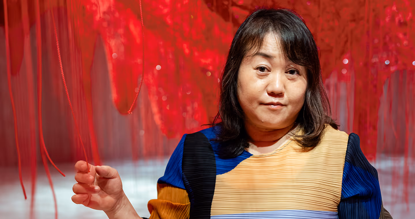 Chiharu Shiota, japanische Künstlerin, 2025 in farbenfrohem Kleid, stehend inmitten ihrer roten Fadeninstallation.