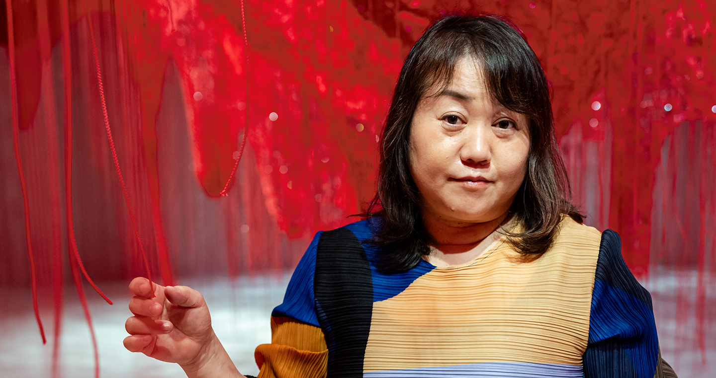 Chiharu Shiota, japanische Künstlerin, 2025 in farbenfrohem Kleid, stehend inmitten ihrer roten Fadeninstallation.