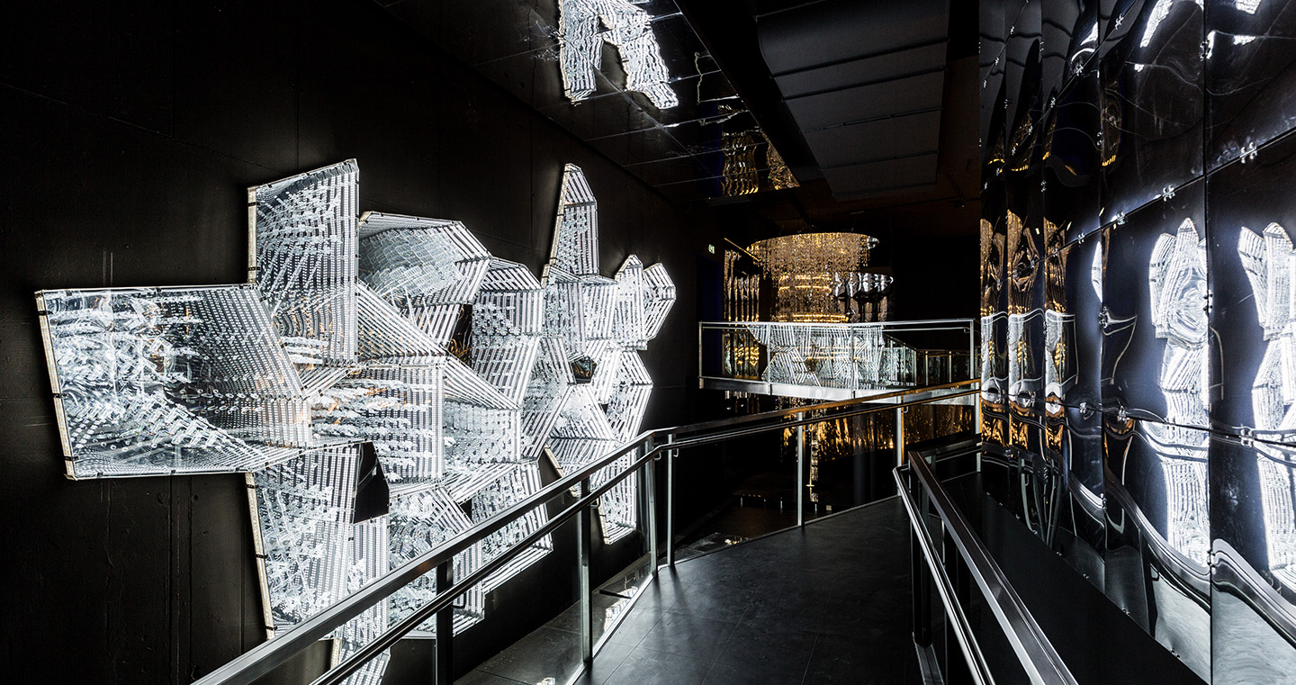Kunstwerk 'Into Lattice Sun': Ein dunkler Raum, durch den ein Steg führt. Links und rechts davon kristalline, urbane Objekte.