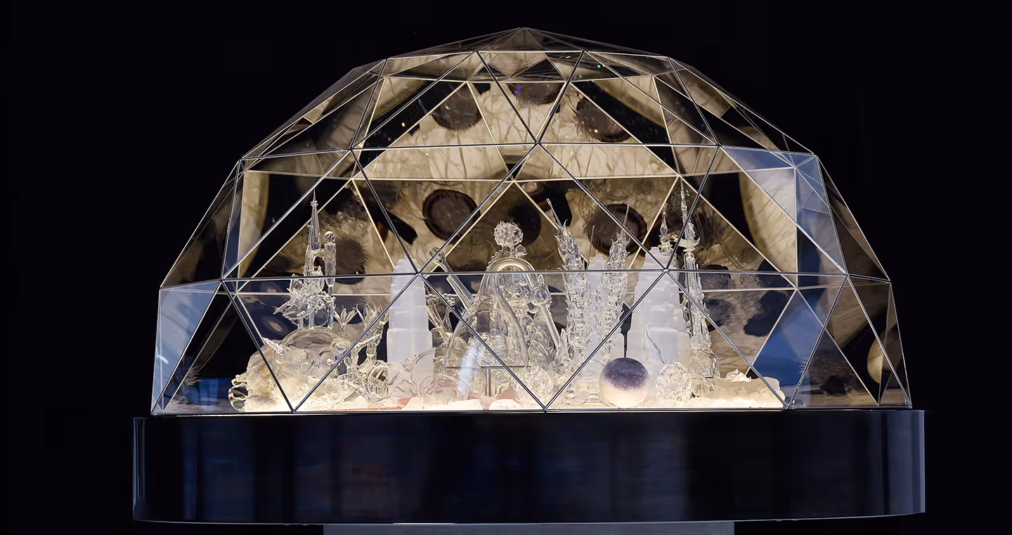Installation 'City of Glass': Eine Miniaturstadt aus gläsernen Gebäuden unter einer Kristallkuppel, kunstvoll beleuchtet.