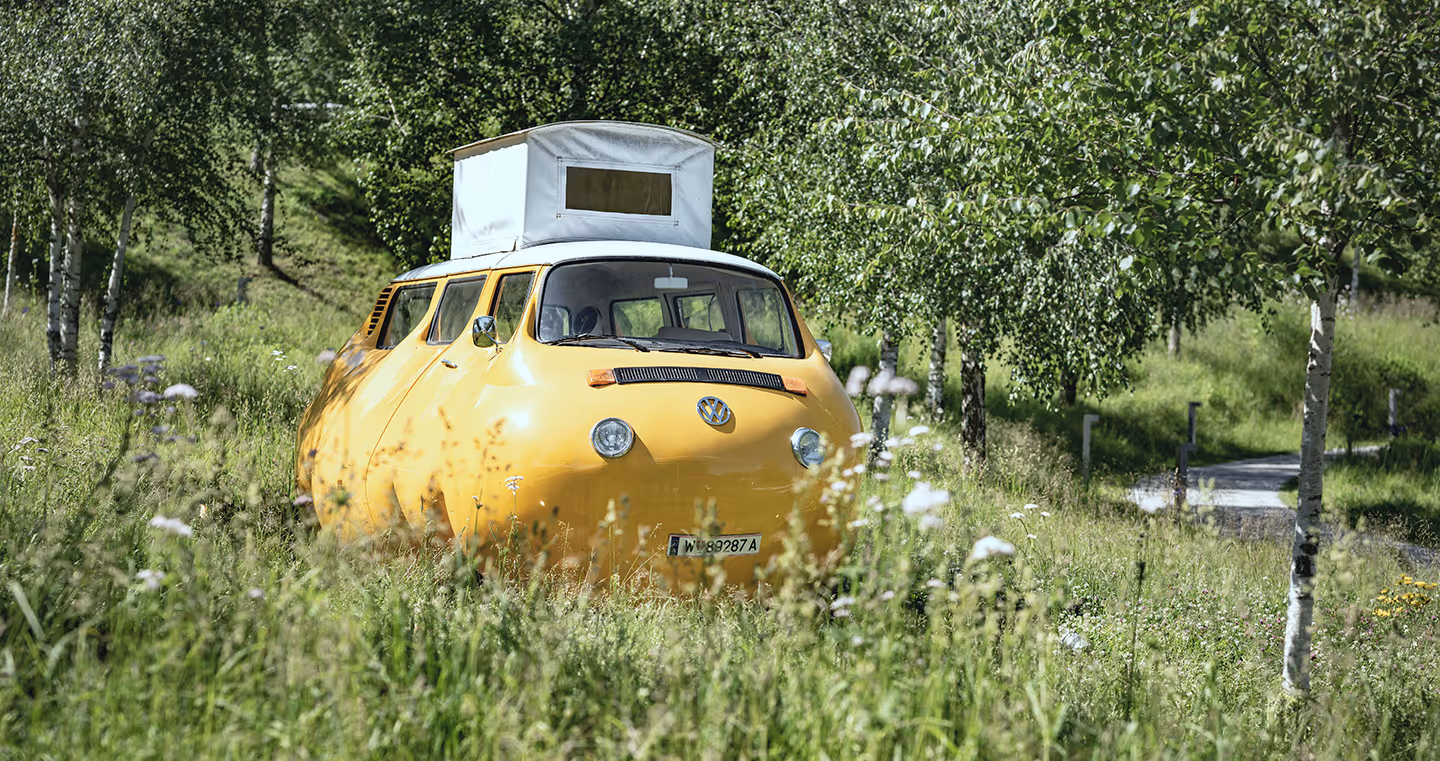 Skulptur 'Dicker Bus': Ein verzerrt wirkender, bauchiger Bus in Grau, humorvoll inszeniert.